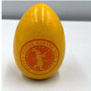 Puede incluir: Un huevo de madera amarillo con un acabado brillante. Una pegatina circular en el huevo dice "WHITE HOUSE EASTER EGG ROLL 2012" y presenta un conejo de dibujos animados. El huevo es un objeto decorativo.