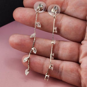May include: A pair of silver-toned drop earrings featuring a slender, textured chain design. Each earring is adorned with multiple marquise-cut clear crystals arranged in a vine-like pattern, finished with a clear plastic earring back.