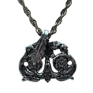 May include: Silver-toned pendant necklace with an ornate, symmetrical design. The pendant features floral and leaf motifs, with a central vertical element and a textured rope chain. The necklace is displayed against a white background.