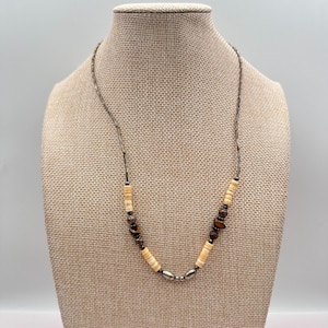 Peut inclure: Un collier avec une chaîne argentée et un pendentif composé de perles crème, marron et argentées. Le collier est présenté sur un mannequin avec un tissu de couleur neutre.
