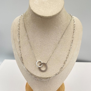 May include: Two silver necklaces displayed on a mannequin. One necklace features a chain with elongated links, while the other has two interlocking circles. The necklaces are made of a shiny, metallic material.