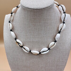May include: A necklace featuring a series of white cowrie shells strung along a thin black cord. The shells are spaced evenly around the circular cord, each secured in place by small knots tied on either side of the individual shell pieces.