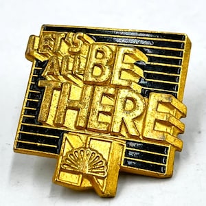 May include: A gold-colored pin with the words "LET'S ALL BE THERE" in raised lettering. The pin has a rectangular shape with black horizontal stripes and a decorative fan design at the bottom. The pin is likely a collectible or promotional item.