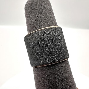 May include: A wide band ring featuring a textured black surface with a matte finish. The ring has a flat profile and a consistent width throughout, showcasing a dark, granular appearance that contrasts with the thin metallic edges visible at the top and bottom.