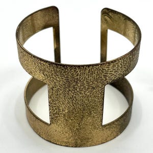 May include: A gold-toned metal cuff bracelet featuring an open-work design with two horizontal bands connected by vertical bars. The surface of the metal has a textured, etched pattern resembling fine, radiating lines across the entire exterior.