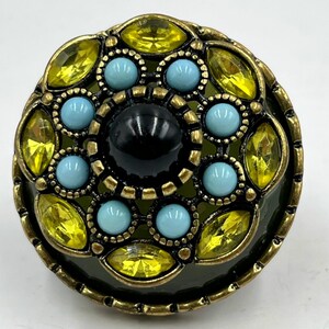 May include: A decorative button with a dark green base and a brass-colored metal frame. The button features yellow and turquoise-colored gemstones and a black center stone. The design is circular and ornate, suitable for embellishing clothing or accessories.