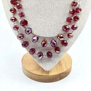 May include: A multi-strand necklace featuring faceted, deep red glass beads strung on a silver-toned metal chain. The beads are round and have a reflective, polished surface that catches the light, creating a layered appearance around the neck.