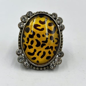 May include: An oval-shaped ring with a yellow and black leopard print center. The ring is framed by a silver-colored metal with small, clear crystals around the edge. A statement piece of jewelry.