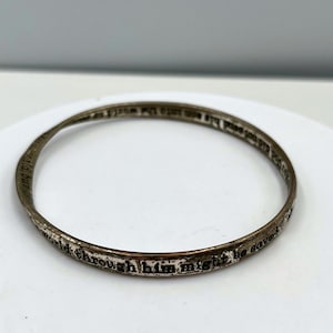 Può includere: Un braccialetto a cerchio argentato con testo inciso. Il testo sembra essere una citazione religiosa. Il braccialetto è circolare e ha un aspetto leggermente usurato.