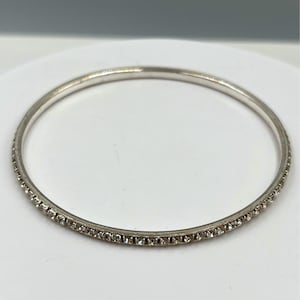 May include: A silver-toned bangle bracelet with a continuous row of small, clear crystals set along one edge. The bracelet is circular and appears to be made of metal. The background is a plain white surface.