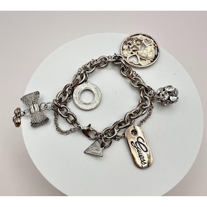 Può includere: Bracciale con ciondoli argentato con diversi ciondoli, tra cui un fiocco, un cerchio, un disegno ad albero, una sfera di cristallo, un triangolo e un'etichetta con "Guess". Il bracciale è su una superficie bianca.
