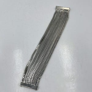 Puede incluir: Una pulsera plateada con múltiples hilos de pequeños eslabones rectangulares. La pulsera tiene un cierre rectangular en cada extremo, y los hilos están conectados al cierre. La joya se muestra sobre una superficie blanca.