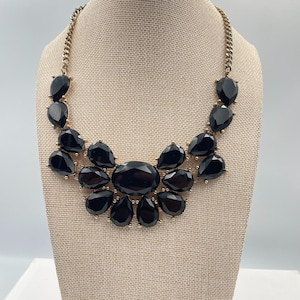 Puede incluir: Un collar llamativo con grandes piedras negras brillantes en forma de lágrima engastadas en metal dorado. El collar tiene una cadena y se exhibe en un maniquí de color neutro.