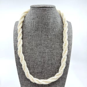 Peut inclure: Un collier composé de plusieurs brins de petites perles blanc cassé en forme de disque, torsadées ensemble pour former un design continu en corde. Les perles sont de taille et de couleur uniformes, créant un aspect texturé et superposé.