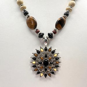 Op de afbeelding: Een ketting met een zilveren hanger in een zonnesterontwerp. De hanger heeft zwarte, bruine en crèmekleurige accenten rond een zwarte ovale steen. De ketting heeft bruine, zwarte en crèmekleurige kralen.