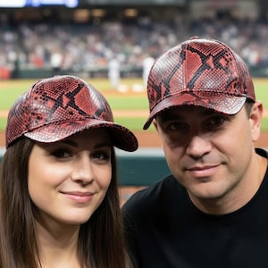 Może przedstawiać: Dwie osoby noszące pasujące czapki bejsbolowe z czerwono-czarnym wzorem skóry węża. Czapki mają zakrzywiony daszek i strukturalny krój. Tło przedstawia stadion baseballowy.