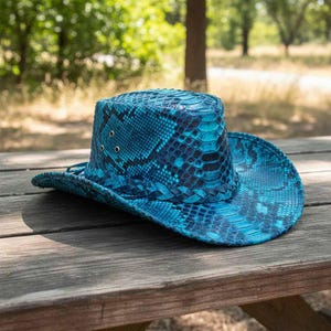 Könnte beinhalten: Ein blauer Cowboyhut mit Schlangenmuster. Der Hut besteht aus einem strukturierten Material und hat eine breite Krempe. Der Hut hat eine dunkelblaue und hellblaue Farbgebung. Der Hut liegt auf einem Picknicktisch aus Holz.