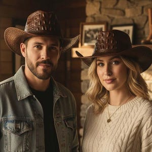 Może przedstawiać: Dwie osoby w brązowych kapeluszach kowbojskich z teksturowanym wzorem przypominającym skórę krokodyla. Kapelusze mają szerokie rondo i zaokrągloną koronę. Mężczyzna ma na sobie dżinsową kurtkę, a kobieta kremowy sweter. Tło obejmuje kominek i oprawione zdjęcia.