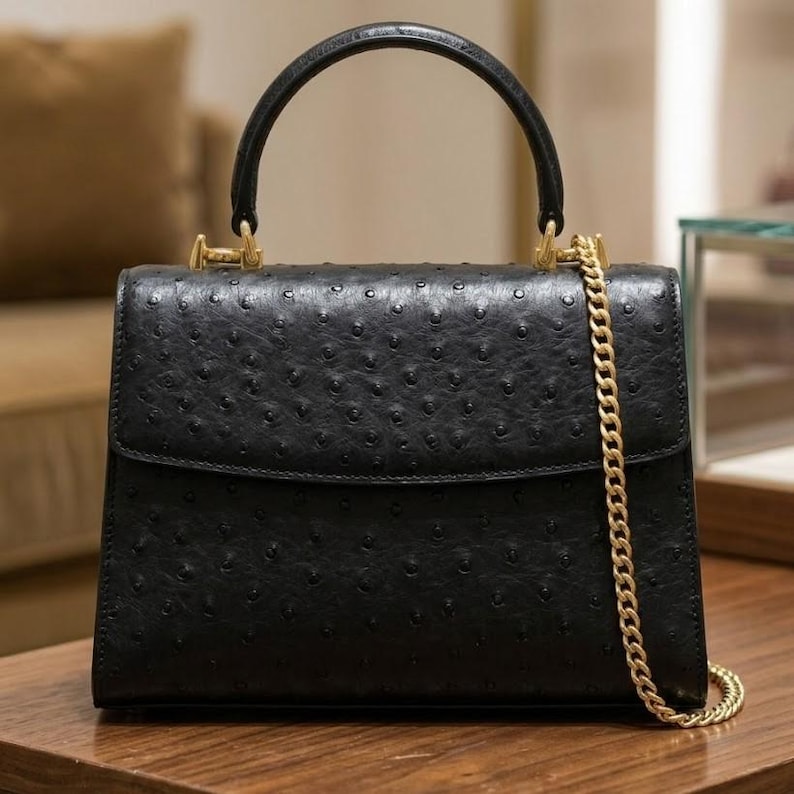 May include: Black leather handbag with a top handle and gold-tone hardware. The bag features a flap closure and a gold chain strap. The leather has a textured, dotted pattern. The bag is sitting on a wooden surface.