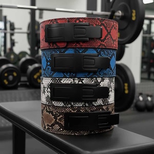 Puede incluir: Una pila de cinturones de halterofilia con diseños de piel de serpiente roja, azul, blanca y marrón. Cada cinturón tiene una hebilla negra y resistente. Los cinturones están apilados en un banco negro en un gimnasio.