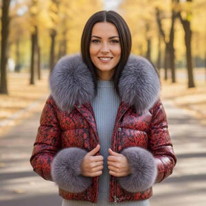 Op de afbeelding: Rode pufferjas met slangenpatroon en grijze bontafwerking rond de kraag en manchetten. De jas is dichtgeritst. De achtergrond is een vage buitenscène met bomen.