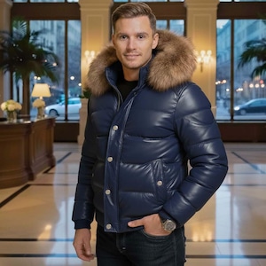 Puede incluir: Una chaqueta acolchada azul marino con capucha ribeteada de piel. La chaqueta tiene un aspecto brillante y acolchado con botones dorados. La persona lleva un reloj y vaqueros azul oscuro. El fondo es un interior bien iluminado.