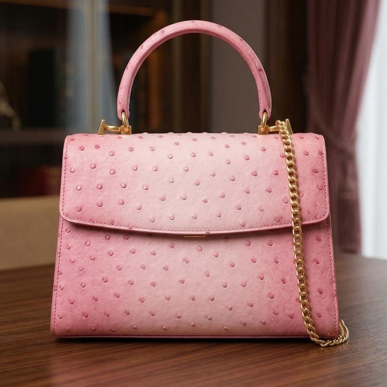 May include: A pink handbag with a top handle and a gold chain strap. The bag has a flap closure and a textured surface with small, evenly spaced dots. The hardware is gold-toned. The bag is sitting on a wooden surface.