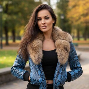 Könnte beinhalten: Eine blaue Jacke mit Schlangenmuster und Pelzkragen. Die Jacke hat einen schwarzen gerippten Saum und Bündchen. Die Person im Bild trägt ein schwarzes Oberteil. Der Hintergrund ist eine verschwommene Szene im Freien mit Bäumen.