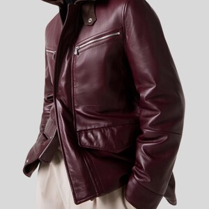 May include: A burgundy leather jacket with a zip-up front, two chest pockets, and two lower pockets. The jacket has a brown suede collar and a black turtleneck underneath. The sleeves are long and the jacket is worn with beige pants.