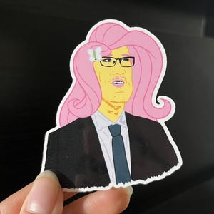 以下が含まれることがあります： ピンクの髪、メガネ、スーツを着た人物の漫画イラストが描かれたステッカー。その人物は黄色い顔をしており、髪には白い花が咲いています。ステッカーは手で持たれています。