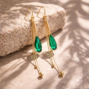 May include: A pair of gold-colored earrings with a dangling design. Each earring features a teardrop-shaped, emerald-green gemstone suspended from a delicate chain. The earrings have a long, flowing style with small gold beads.
