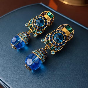 May include: A pair of ornate earrings with a gold-tone setting. Each earring features a large, faceted blue crystal teardrop pendant. The earrings are adorned with smaller blue and green crystals, creating a luxurious and eye-catching design.