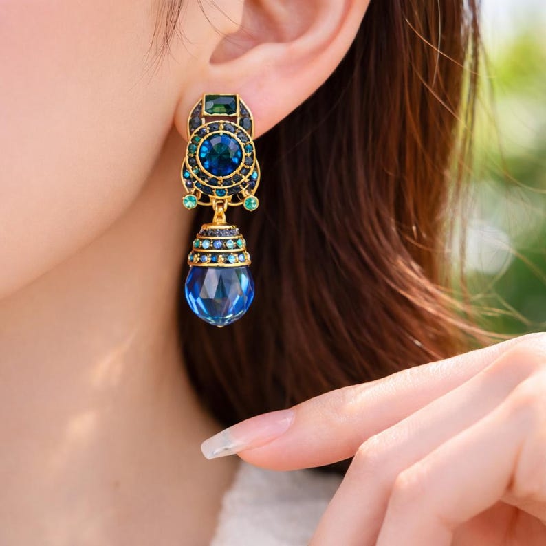 May include: Gold-toned drop earrings with blue and green gemstones. The earrings feature a large, faceted blue teardrop-shaped stone at the bottom, with intricate detailing and smaller stones in a circular design. The earrings are worn on an ear.