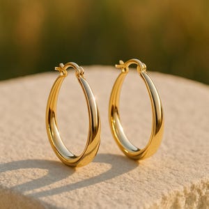 May include: A pair of gold-colored oval hoop earrings. The earrings are smooth and shiny, reflecting light. The hoops are a classic design, suitable for everyday wear or special occasions. The earrings are displayed on a neutral-colored surface.