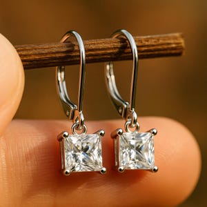 May include: A pair of silver leverback earrings with square, clear gemstones. The earrings are held up by a wooden stick, with a blurred background. The gemstones are set in a square shape and appear to be sparkling.
