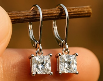 May include: A pair of silver leverback earrings with square, clear gemstones. The earrings are held up by a wooden stick, with a blurred background. The gemstones are set in a square shape and appear to be sparkling.