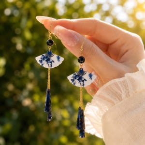 May include: Dangle earrings featuring white fan-shaped charms with blue floral patterns, suspended from gold-tone hooks. Each earring includes a black bead, a gold chain, and a dark blue marbled acrylic pendant at the base of the design.