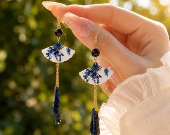 May include: Dangle earrings featuring white fan-shaped charms with blue floral patterns, suspended from gold-tone hooks. Each earring includes a black bead, a gold chain, and a dark blue marbled acrylic pendant at the base of the design.
