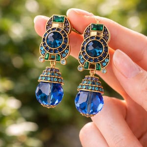 May include: A pair of ornate earrings with a gold-tone setting. Each earring features a large, round, deep blue crystal at the center, surrounded by smaller blue and green crystals. The earrings have a tiered design with a large, teardrop-shaped blue crystal at the bottom.
