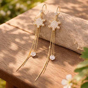May include: A pair of gold-colored dangle earrings. Each earring features a snowflake-shaped design encrusted with small, clear crystals. Below the snowflake, three gold chains hang, ending with a round, clear crystal. The earrings are displayed on a wooden surface.