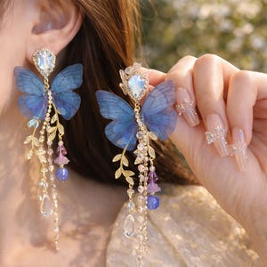 May include: Elegant butterfly earrings with blue wings and sparkling crystal accents. The earrings feature a gold-tone metal design with dangling chains, purple beads, and clear teardrop-shaped crystals. The earrings are a beautiful accessory.