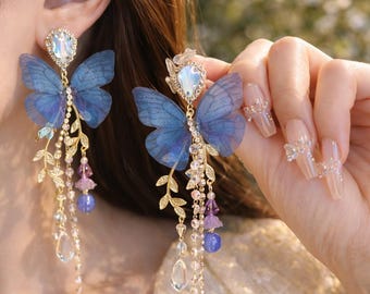May include: Elegant butterfly earrings with blue wings and sparkling crystal accents. The earrings feature a gold-tone metal design with dangling chains, purple beads, and clear teardrop-shaped crystals. The earrings are a beautiful accessory.