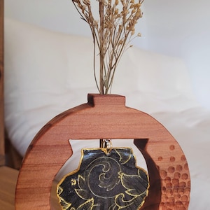 Op de afbeelding: Een cirkelvormige houten vaas met een getextureerde rechterkant, gevuld met gedroogde beige bloemen. Een zwarte schelphanger met goudkleurige, wervelende bladpatronen hangt aan een kleine metalen ring in de centrale opening van het houten frame.