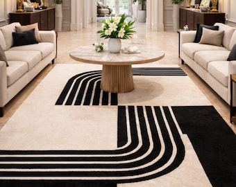 May include: A cream-colored area rug with a bold black geometric design. The rug features curved and straight lines, creating a modern, abstract pattern. The rug is placed in a spacious living room setting with cream-colored sofas and a round coffee table.