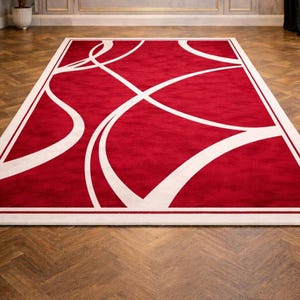 May include: A rectangular red rug with a white border and abstract white curved lines. The rug is placed on a wooden floor with a herringbone pattern. The room has a neutral color palette with gold accents and a potted plant.