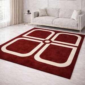 May include: A rectangular area rug with a geometric design. The rug is primarily a deep red color, with a large, off-white, four-leaf clover-like pattern in the center. The rug is placed in a living room setting.