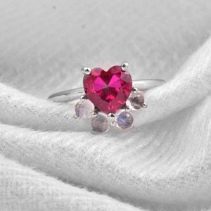 May include: A silver ring with a heart-shaped, deep red gemstone at its centre. The heart is encircled by smaller, round, iridescent stones. The ring is set against a white, textured backdrop, accentuating the jewellery's design.