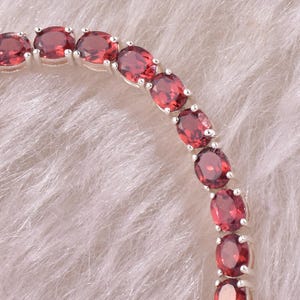 May include: A silver bracelet featuring a series of oval-cut, deep red gemstones. The gemstones are set closely together, creating a continuous line of color. The bracelet is displayed on a soft, white, textured surface.