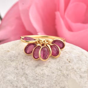 May include: A gold ring featuring a cluster of six oval-shaped, deep pink gemstones. The ring is set against a soft, white surface with a blurred pink flower in the background. The gemstones are suspended from the ring's band.
