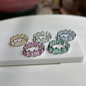 May include: Five silver rings, each featuring a continuous band of oval-shaped gemstones in various colours including yellow, light green, pink, dark green, and blue. The stones are set in a simple metal frame, creating a uniform, circular design.